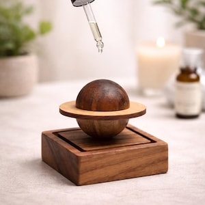 Peut inclure: Diffuseur d'huiles essentielles en bois avec un design en forme de planète. Le diffuseur a une sphère marron foncé, un anneau marron clair et repose sur une base carrée. Une pipette dépose de l'huile sur la sphère. En arrière-plan, une bougie et une petite bouteille.
