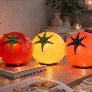 May include: Three tomato-shaped lamps in red, yellow, and orange. Each lamp has a green star-shaped top and sits on a wooden surface. The lamps are lit, creating a warm glow, and are placed on a table.