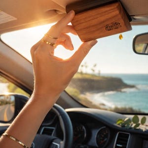 Peut inclure: Un diffuseur de parfum en bois pour voiture avec le mot "drift" imprimé dessus. Le diffuseur est rectangulaire et est tenu dans une voiture. Une goutte de liquide ambré pend en bas. L'arrière-plan montre une journée ensoleillée avec l'océan au loin.