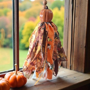May include: A decorative hanging ornament with a ceramic pumpkin and fabric strips in autumnal colours. The ornament is suspended by a metal hook and wooden beads. Two small pumpkins sit on a wooden windowsill, near a window.