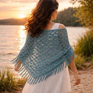 May include: A blue crocheted shawl with fringe. The shawl is worn over a white dress. The image is taken outdoors with a lake and sunset in the background. The shawl has a delicate open weave pattern.