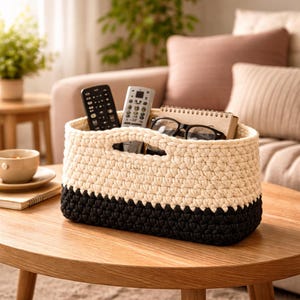 May include: A two-tone, hand-crocheted basket in cream and black, with a rectangular shape and two handle openings. The basket holds two remote controls, a pair of glasses, and a notebook. It sits on a wooden table in a living room setting.