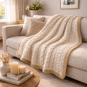 May include: Cream cable knit throw blanket with a tan border draped over a beige sofa. A matching pillow rests against the back of the sofa. The blanket features a textured, woven design, creating a cozy and inviting aesthetic.