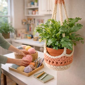 May include: A hanging plant holder with a terracotta pot filled with green leafy plants. The holder is made of cream and peach-colored crochet yarn. A person is arranging yarn balls in a basket and tray on a white table.
