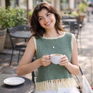 May include: A sleeveless green knit top with cream-colored trim and fringe. The top is paired with white pants. A woman is holding a white coffee cup and saucer. She is wearing a gold necklace with a pearl pendant.