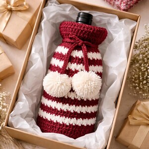 May include: A red and white striped wine bottle cozy with two cream-colored pom-poms. The cozy is inside a cardboard box with white tissue paper. The bottle is partially visible, and the cozy is tied with a matching red string.