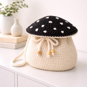 May include: A cream-colored crochet bag shaped like a mushroom, with a black top dotted with white spots. The bag has a drawstring closure with wooden beads and a cream-colored strap.