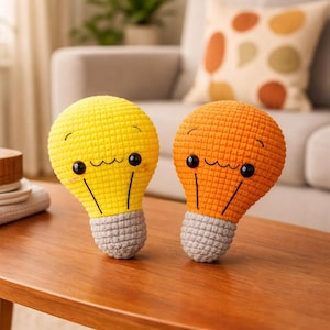 May include: Two crocheted light bulb plushies, one yellow and one orange, with smiling faces and gray bases. The bulbs have black eyes and black lines for the light filaments. They are sitting on a wooden table.