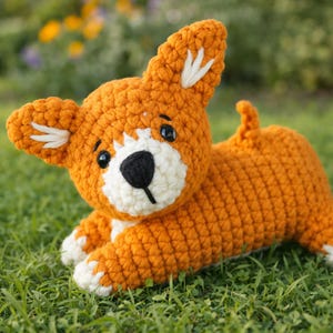 May include: A handmade orange crochet dog toy with white accents on the ears, paws, and face. The dog has black button eyes and a black nose. The toy is lying on green grass.
