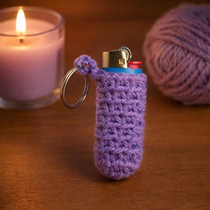 May include: A purple crocheted lighter cozy with a silver keyring attached. The lighter has a blue base and gold metal top with a red button. A lit candle and a ball of purple yarn are in the background. The cozy is handmade.