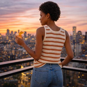 Puede incluir: Un top de crochet a rayas crema y naranja. El top sin mangas se combina con unos vaqueros azules. La persona sostiene un cóctel con una rodaja de naranja, con vistas al horizonte de una ciudad al atardecer. El top es una prenda informal, hecha a mano.
