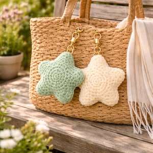 Könnte beinhalten: Zwei gehäkelte sternförmige Schlüsselanhänger, einer hellgrün und einer cremefarben, hängen an goldfarbenen Verschlüssen. Sie befinden sich vor einer geflochtenen Stroh-Tragetasche. Ein gestreifter Schal hängt auf der rechten Seite der Tasche, mit verschwommenem Grün im Hintergrund.