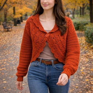 May include: An orange crochet cardigan with a scalloped collar and a single button closure. The long-sleeved sweater is paired with blue jeans and a brown belt. The background shows a park with autumn foliage.