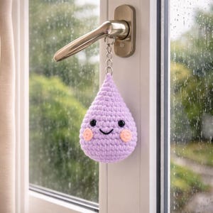 以下が含まれることがあります： 黒い目と桃色の頬を持つ、手作りのラベンダー色の編み物雨滴キーホルダー。ドアノブに付いたシルバーチェーンから吊り下げられています。背景には雨の降る窓と、ぼやけた緑色の屋外の景色が写っています。