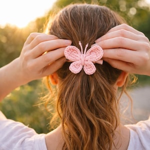 May include: A light pink crochet butterfly hair clip. The butterfly is centered in the image, attached to a ponytail. The hair clip is made of a soft, textured yarn. The background is blurred, with sunlight filtering through the trees.
