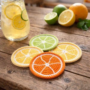 Könnte beinhalten: Handgefertigte Häkeluntersetzer in Form von Zitrusfruchtscheiben: Orange, Zitrone und Limette. Die Untersetzer liegen auf einer rustikalen Holzoberfläche, mit einer Limonadenkanne und frischen Früchten im Hintergrund.