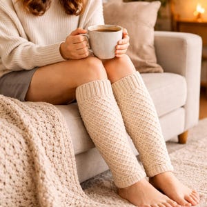 May include: Cream-colored knitted leg warmers with a diamond pattern. The leg warmers are worn on the legs of a person sitting on a couch. A matching knitted blanket is draped over the couch. A person is holding a mug of coffee.