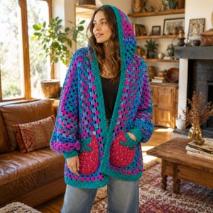 Pode incluir: Um cardigan de crochê com capuz colorido em tons de azul, rosa e verde-azulado. O suéter de frente aberta apresenta um padrão de quadrados da vovó, bolsos grandes e um capuz. Os bolsos são de cor rosa brilhante. As mangas são longas.