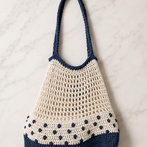 Pode incluir: Uma bolsa de crochê com corpo de malha aberta na cor creme e base sólida azul-marinho. A bolsa inclui uma alça azul-marinho combinando e uma borda decorativa na parte inferior adornada com pequenos pontos em relevo azul-marinho.