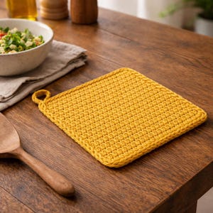 Pode incluir: Um pegador quadrado amarelo de crochê com uma alça para pendurar. O pegador é feito de fio texturizado e está sobre uma mesa de madeira. Uma colher de pau e uma tigela de salada também são visíveis.