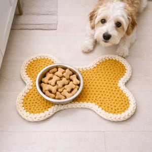 Peut inclure: Un tapis de nourriture pour chien en crochet jaune moutarde en forme d'os, avec une bordure crème. Un bol en acier inoxydable rempli de biscuits pour chiens est au centre. Un petit chien duveteux avec des poils bruns et blancs est en arrière-plan.