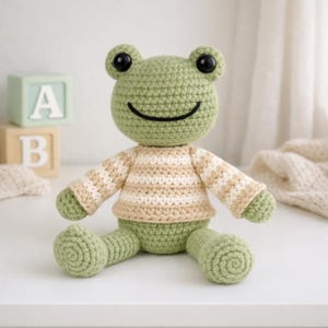 Op de afbeelding: Een handgemaakte gehaakte kikkerpop in groen, beige en wit. De kikker heeft grote zwarte ogen en een lachende mond. Hij draagt een gestreepte trui. Een charmant, zacht speelgoed.