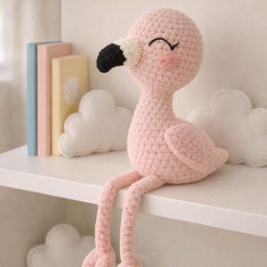 Pode incluir: Uma boneca de flamingo rosa de crochê feita à mão com um bico preto e branco, sentada em uma prateleira branca. O flamingo tem um rubor em forma de coração e olhos fechados. Almofadas decorativas em forma de nuvem e livros coloridos ao fundo.