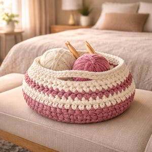 May include: A cream and mauve crochet basket, holding balls of yarn and wooden crochet hooks. The basket is handmade with a handle, ideal for storing craft supplies. The yarn balls are cream and mauve, perfect for knitting or crochet.