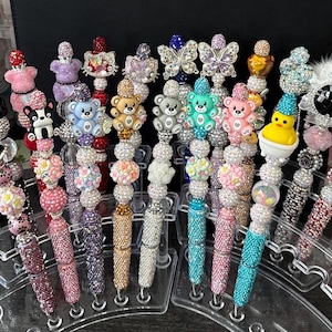 May include: A selection of decorative pens with a variety of adornments. The pens are adorned with colourful rhinestones, beads, and charms, including teddy bears, butterflies, and flowers. The pens are displayed on a clear acrylic stand.