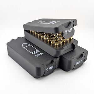 May include: Black 9mm ammunition storage cases stacked on a white surface. Each case has a hinged lid with a white outline of a bullet and the text "9MM". One case is open, revealing rows of brass-colored bullets.
