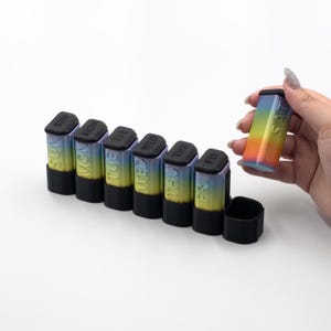 May include: A seven-day pill organizer featuring individual vertical containers with black caps and bases. Each container has a gradient finish transitioning from blue to yellow to orange, with the days of the week embossed on the side.