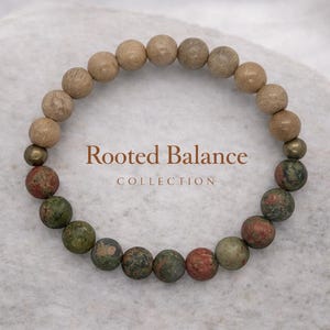 Könnte beinhalten: Ein Armband mit hellbraunen und grün-roten Steinperlen. Das Armband enthält einige messingfarbene Perlen. Der Text "Rooted Balance COLLECTION" ist in brauner Schrift dargestellt.