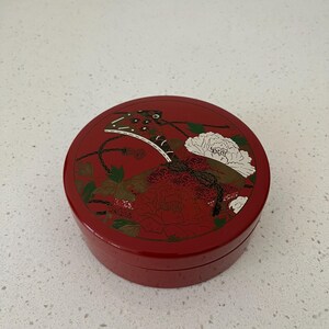May include: A round, red lacquered box with a lid featuring a floral design and a stylized insect. The design includes white and green accents on a red background. The box is likely used for storage or display.