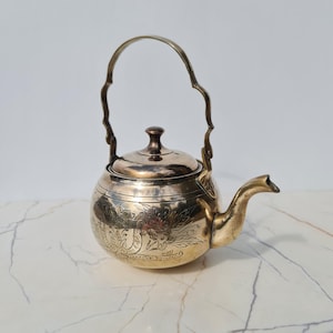 May include: A metallic brass teapot featuring a rounded body with intricate floral engravings, a curved spout, and a tall, wavy-edged handle. The lid is domed with a small central knob, and the surface displays a polished, reflective finish.