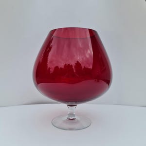 May include: A large, deep red glass vessel with a wide, rounded bowl shape that tapers towards the top opening. The piece features a clear, short stem and a circular base, providing a stable foundation for the translucent, coloured glass body.