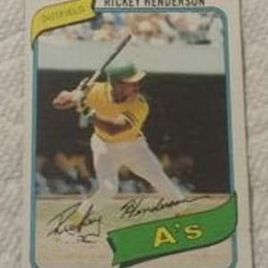 May include: A vintage baseball card featuring Rickey Henderson in an Oakland A's uniform. The card has a blue border and includes the text "Rickey Henderson" at the top. Henderson is shown mid-swing, wearing a yellow and green uniform.