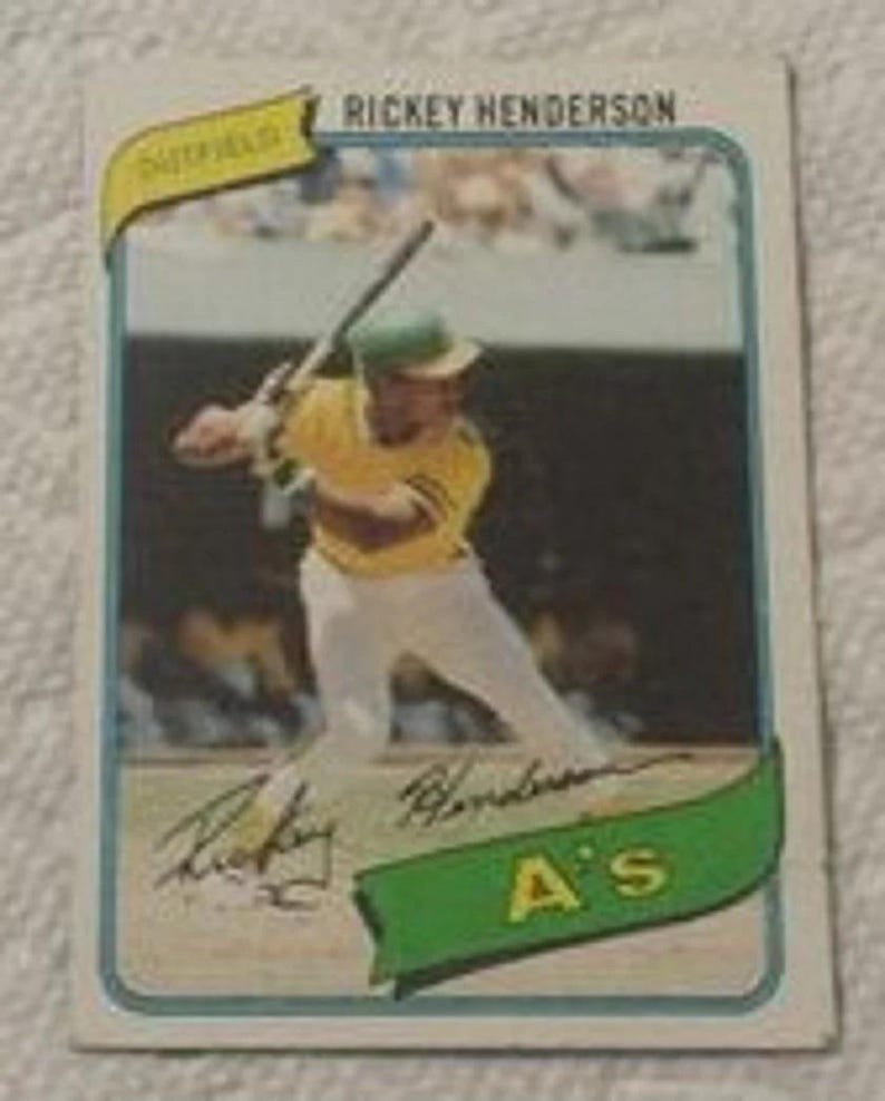 May include: A vintage baseball card featuring Rickey Henderson in an Oakland A's uniform. The card has a blue border and includes the text "Rickey Henderson" at the top. Henderson is shown mid-swing, wearing a yellow and green uniform.