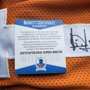 May include: Close-up of an orange mesh jersey with a white patch and a Beckett Certificate of Authenticity. The certificate has text and a blue and white logo. The jersey has a signature.