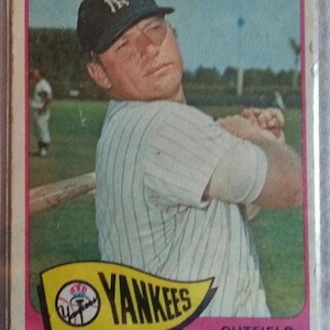May include: Vintage baseball card featuring Mickey Mantle of the New York Yankees. The card shows Mantle in a pinstriped uniform and a Yankees cap. The card has a pink border and the Yankees logo.