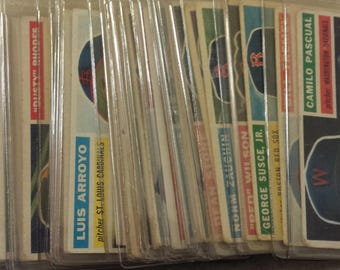 May include: A collection of vintage baseball cards, each encased in a protective sleeve. The cards feature player portraits and team logos, with names like Don Mossi and Camilo Pascual. The cards are in various colors.