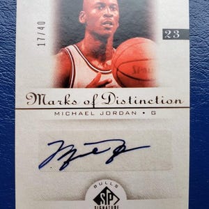May include: A Michael Jordan basketball card from the "Marks of Distinction" series. The card features a portrait of Jordan holding a basketball, with his signature and the text "SP Signature Edition". The card is numbered 17/40 and has the number 23.