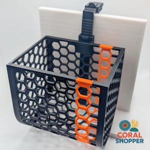 May include: A black and orange plastic basket with a hexagonal grid pattern, featuring a vertical orange accent strip on the front. The basket is attached to a black adjustable mounting bracket and is positioned against a flat white square panel.