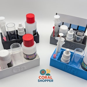 May include: Four aquarium test kits in black, white, and blue trays. Each kit contains bottles, syringes, and vials. The trays are labeled with "Ca", "Mg", and "04". The bottles have red caps. The Coral Shopper logo is in the bottom right.