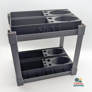 May include: A gray, two-tiered organizer with compartments and circular openings. The top level has "NO3" text, and the bottom level has "Ca" text. The Coral Shopper logo is in the bottom right corner.