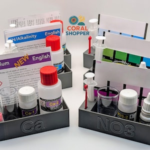 May include: Gray plastic trays containing aquarium water testing kits. Each kit includes bottles with red caps, syringes, test tubes, and color charts. The trays are labeled with "Ca" and "NO3" and include English instructions. The Coral Shopper logo is visible.