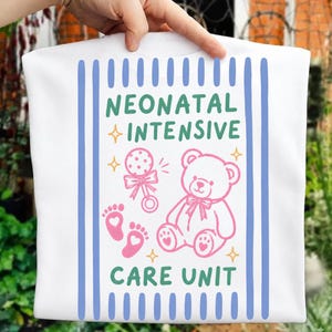 May include: A white t-shirt featuring a graphic with the text NEONATAL INTENSIVE CARE UNIT in green. The design includes a pink teddy bear, a pink rattle, and two pink baby footprints with heart centers, all framed by vertical blue stripes.