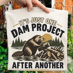 May include: A cream-colored t-shirt featuring a graphic of a beaver building a dam with logs in water. The text reads IT'S JUST ONE DAM PROJECT AFTER ANOTHER in black and tan lettering, set against a backdrop of pine trees and a sun.