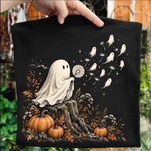May include: A black t-shirt featuring a graphic of a white ghost sitting on a tree stump, blowing on a dandelion. The dandelion seeds transform into smaller ghosts floating away. Two orange pumpkins and autumn foliage surround the tree stump.