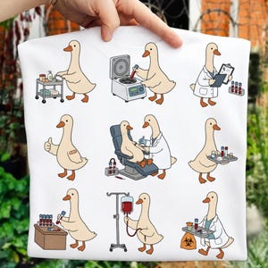 May include: A white t-shirt featuring a repeating pattern of cartoon ducks in various medical settings, including a duck with a medical cart, a duck using a centrifuge, a duck with a clipboard, a duck with a bandage, and a duck receiving an IV.
