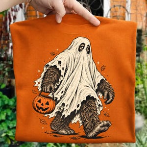 May include: An orange t-shirt featuring a graphic illustration of a brown, furry creature wearing a white ghost sheet. The creature is holding an orange jack-o'-lantern pumpkin bucket, surrounded by small falling autumn leaves and star shapes.